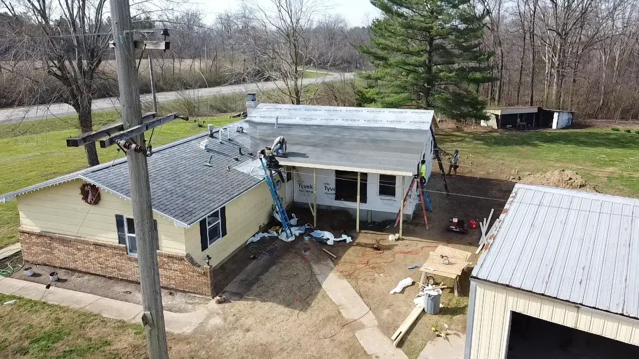 Aerial view of a small house with a white roof, yard, and nearby shed in a rural setting.