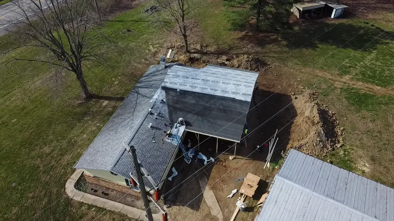 Aerial view of a house with a gray roof, fenced yard, and scattered yard items beside a white structure.