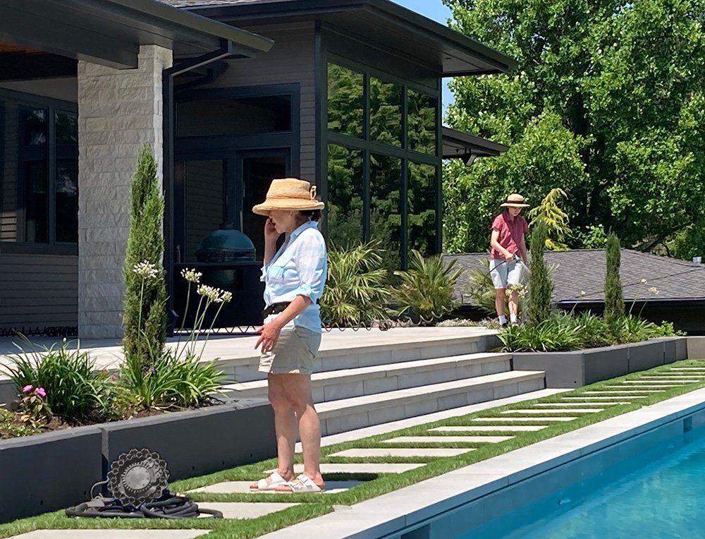 Two people by a modern poolside patio, one standing on stepping stones and one near the steps, on a sunny day