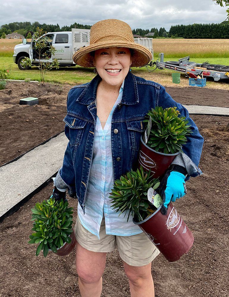 Smiling person in a straw hat carrying potted plants in a field, with a truck in the background