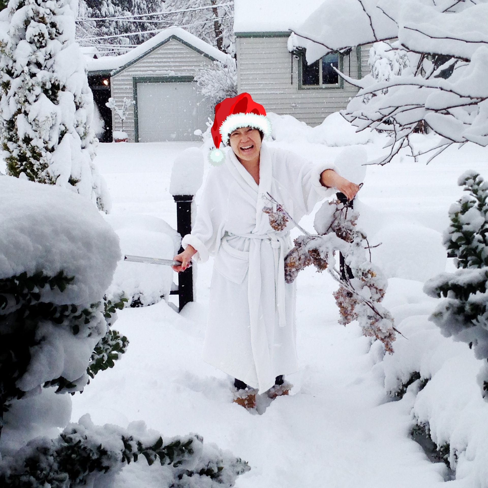 Person in a Santa hat standing in deep snow between snow-covered bushes and houses, smiling.
