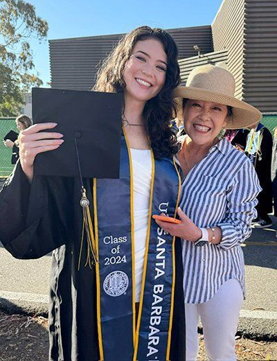 Two women smile at a graduation, one in cap and gown holding a diploma cover outdoors.