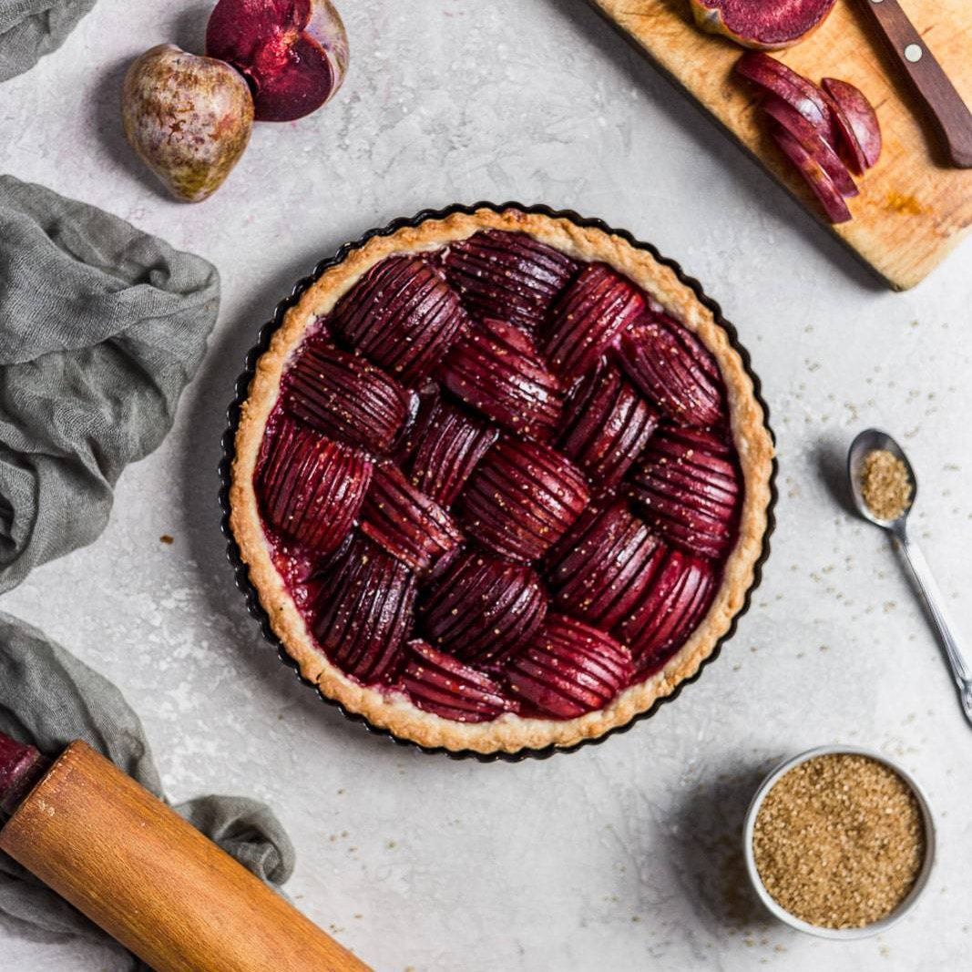 Round plum tart with lattice top on a gray surface, surrounded by plums, spoon, and baking tools.