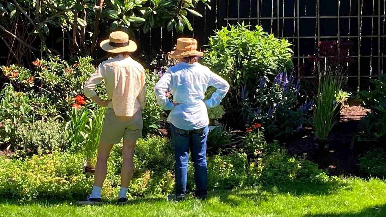 Two people in straw hats standing in a sunny garden, facing a lush flower bed and greenery.