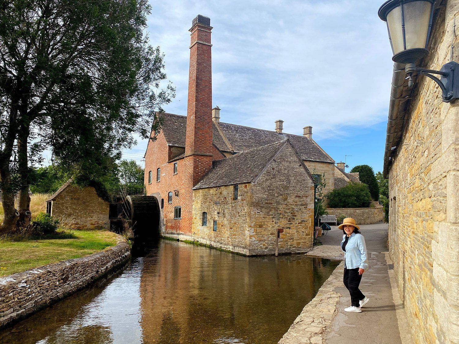 Canal beside old brick building and chimney; person walking along stone path under a cloudy sky
