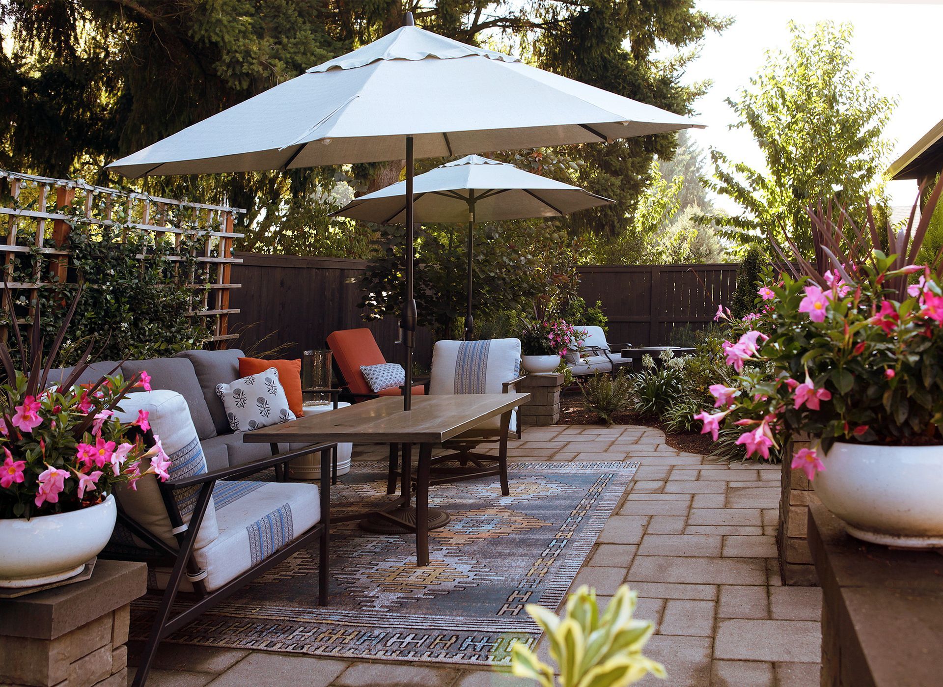 Patio seating area with white umbrellas, cushioned chairs, wooden table, and potted flowers in a garden.