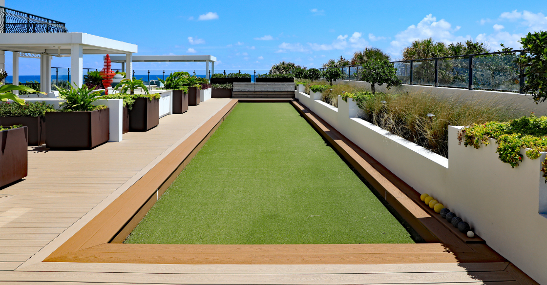 Modern rooftop garden with a long green lawn, planters, and white benches under a bright blue sky