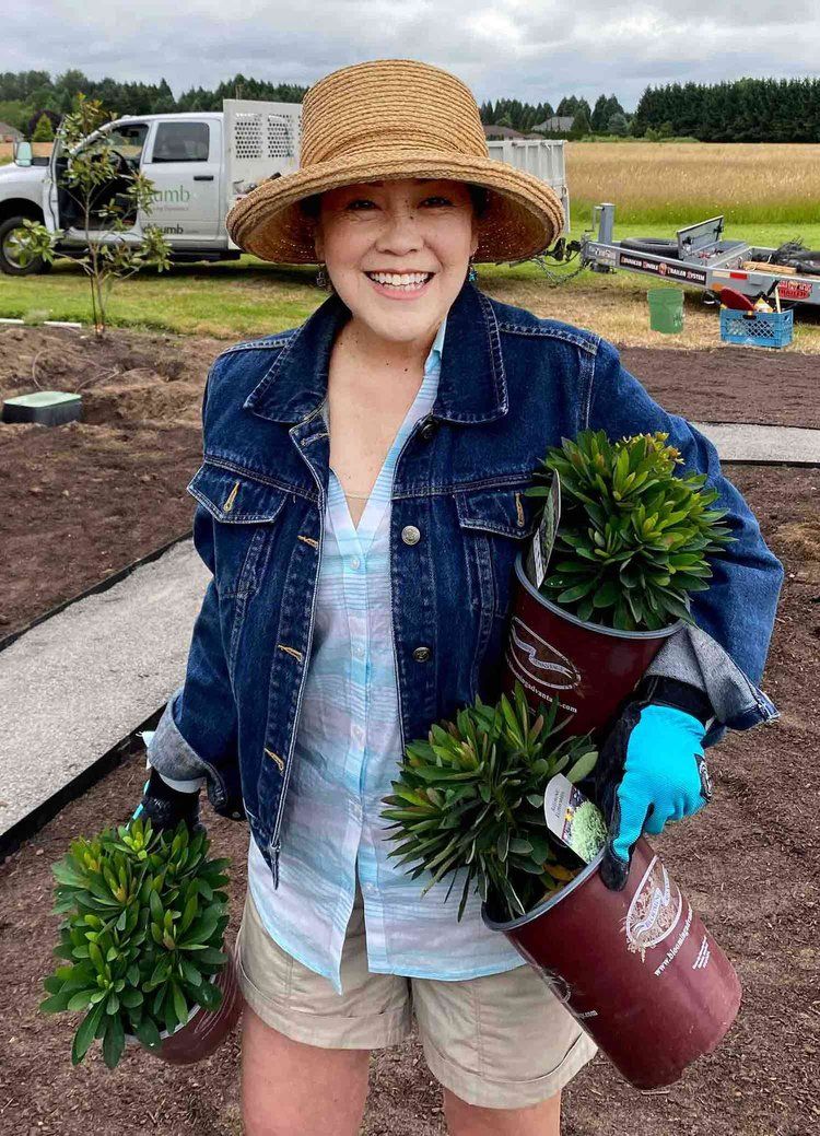 Person in straw hat and denim jacket holding potted plants in a garden field, smiling.