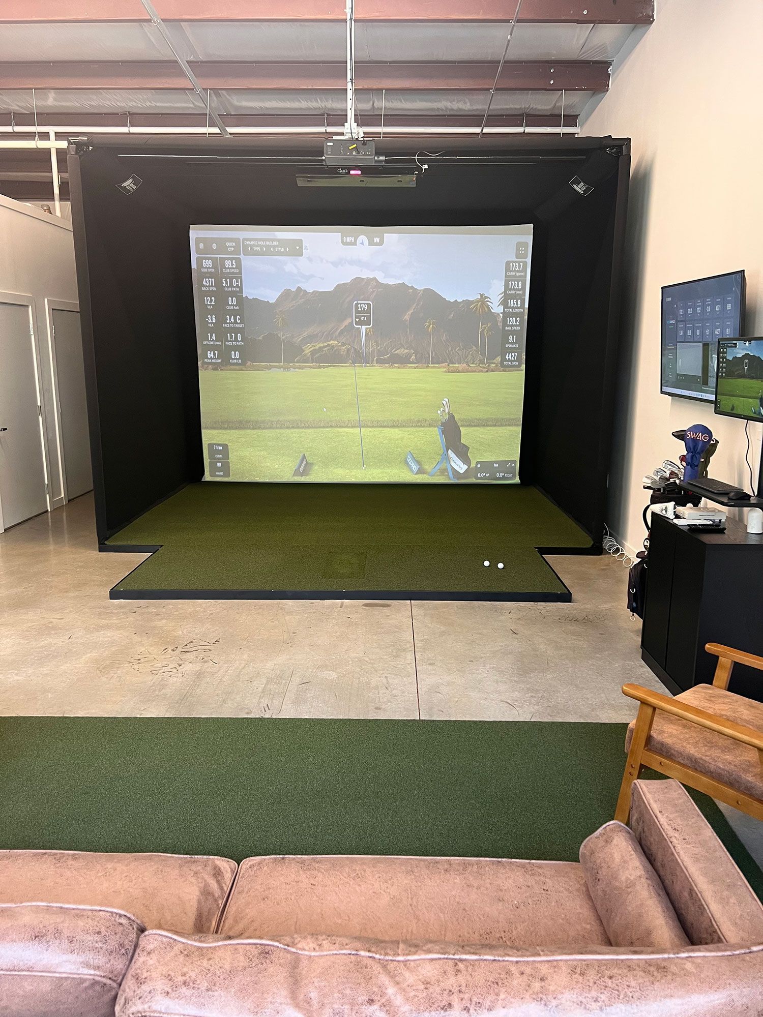Indoor golf simulator bay with large projection screen, turf mat, and seating area.