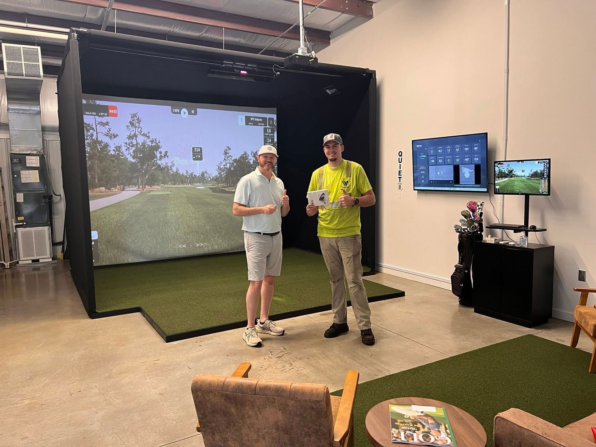 Two people standing indoors beside a golf simulator screen and monitors in a game room