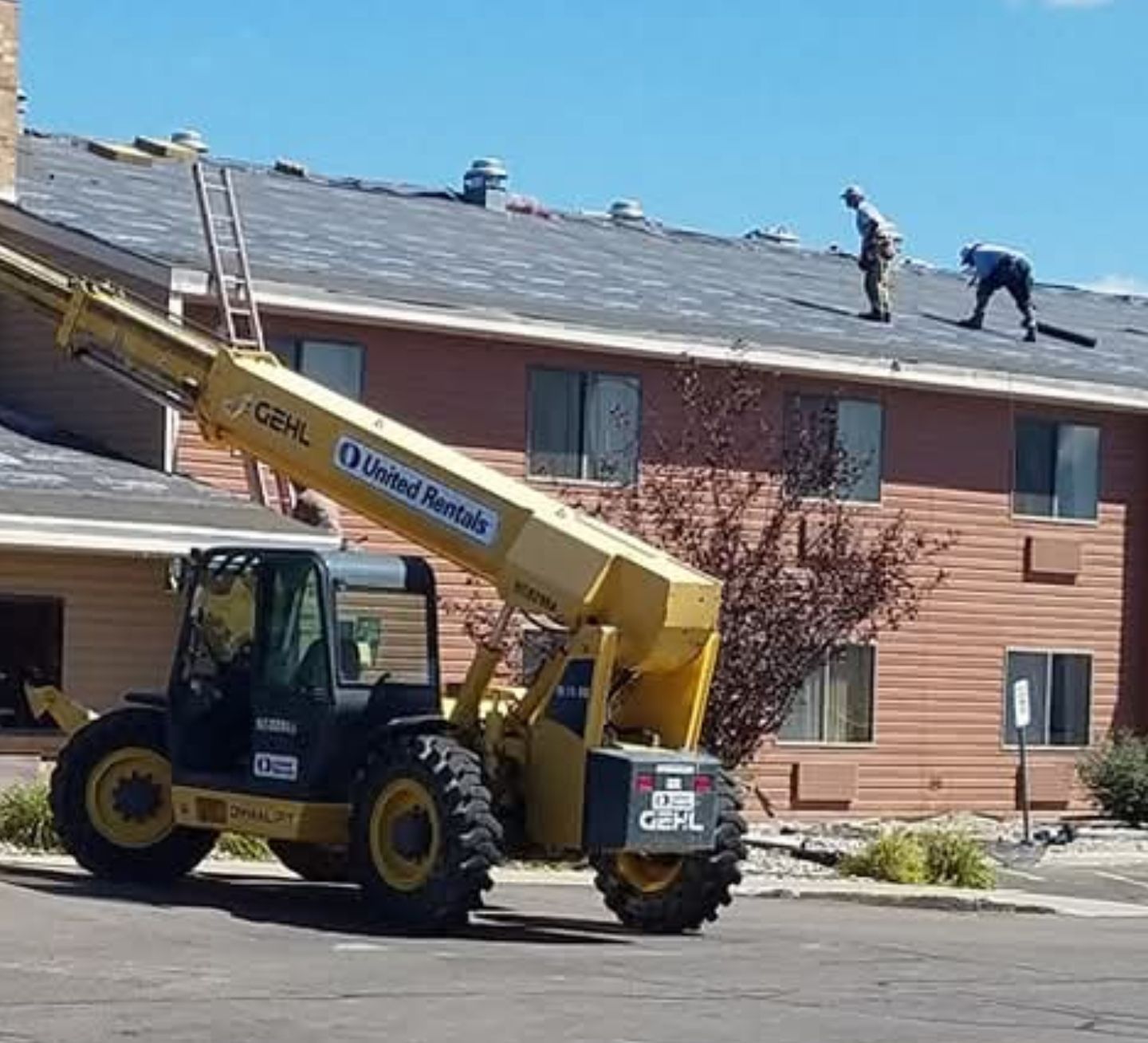Construction crew repairing a roof with a yellow crane truck beside a tan apartment building