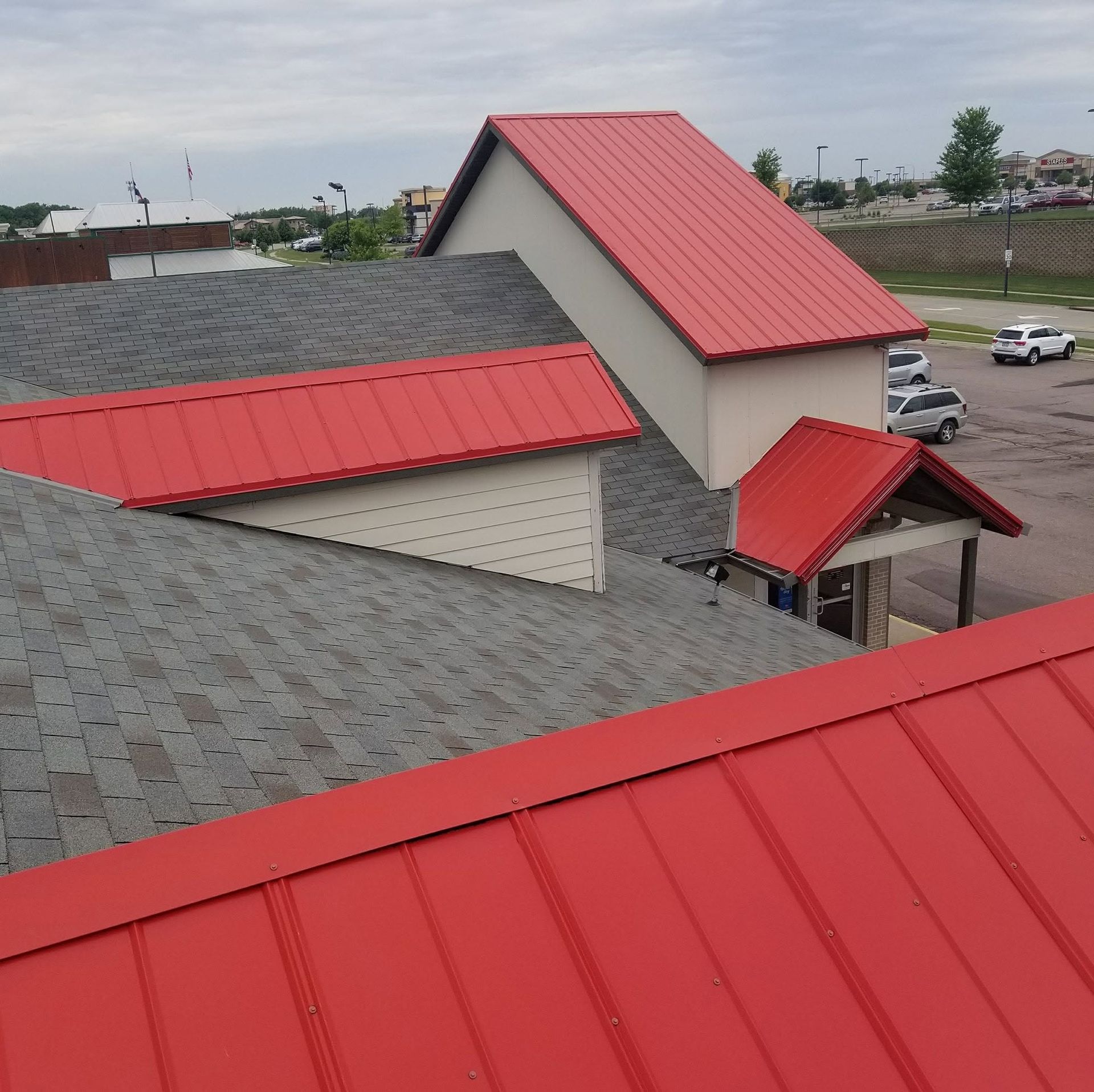 Red metal roofs on a beige building complex under a cloudy sky, with a parking lot nearby