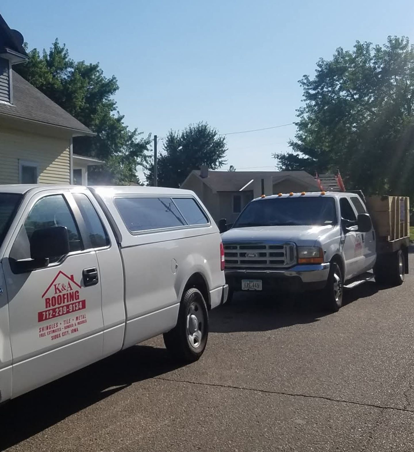 Two work trucks parked on a sunny residential street, one white and one silver with a trailer.