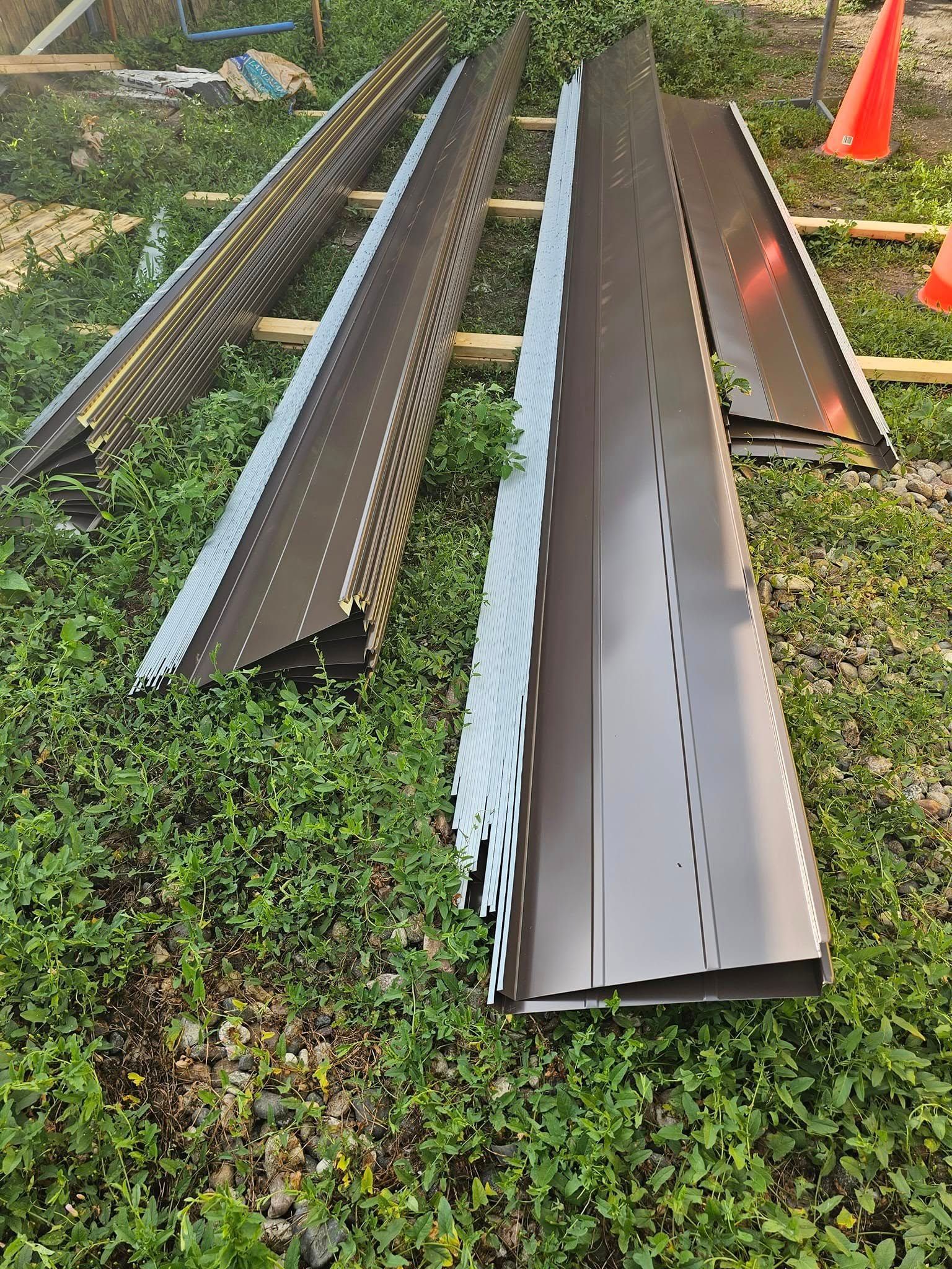 Long metal roofing panels laid on grass near an orange traffic cone outdoors