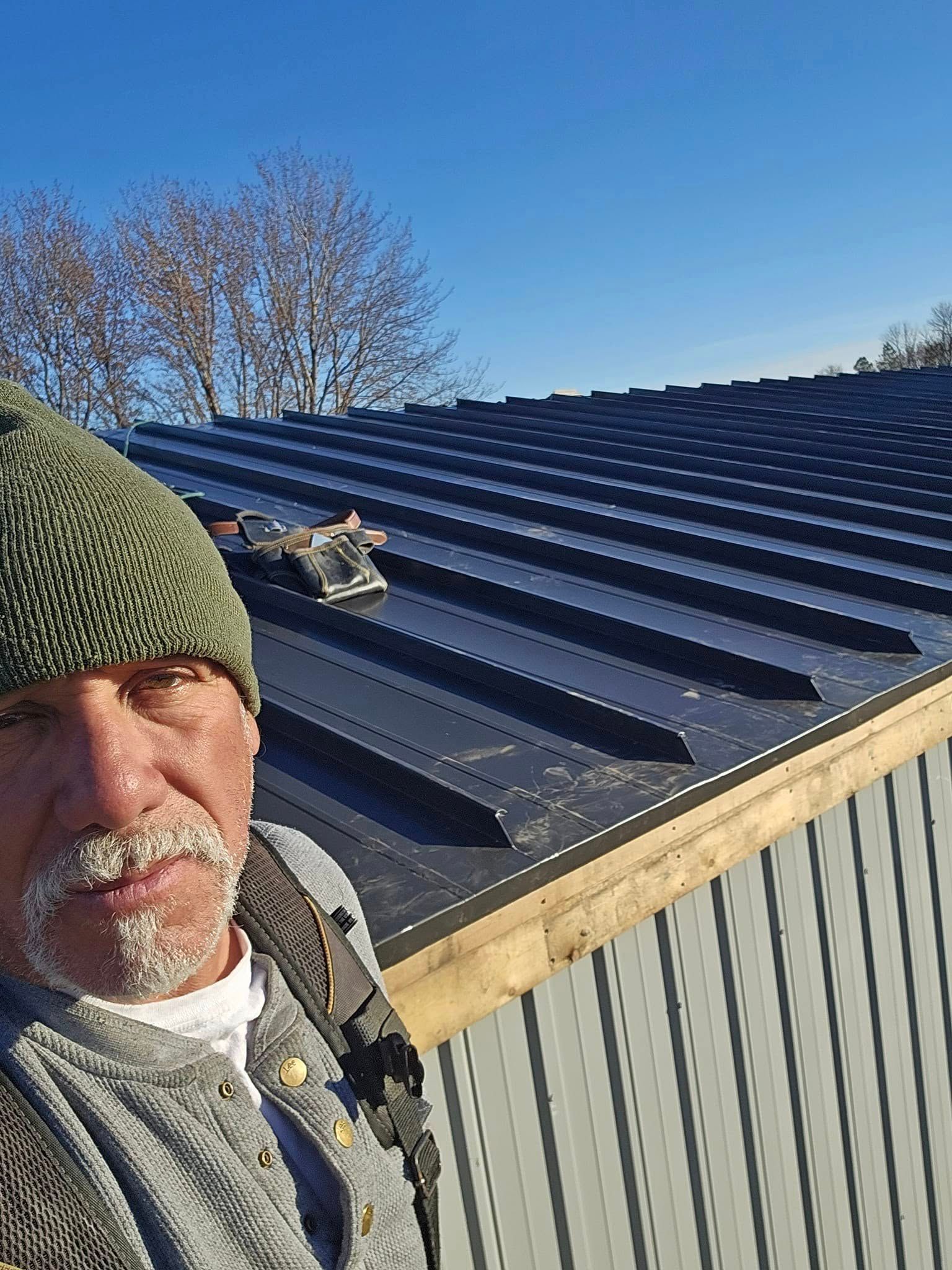 Worker on a corrugated metal roof, wearing a green knit cap and safety harness, under a blue sky.