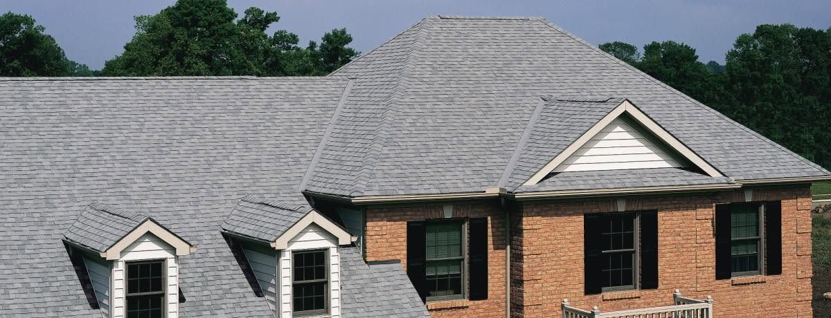 Brick house with gray shingled roof and dormer windows against a green tree line