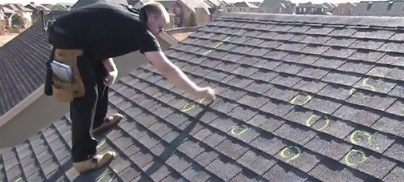 Worker applying adhesive to roof shingles while kneeling on a sloped roof.