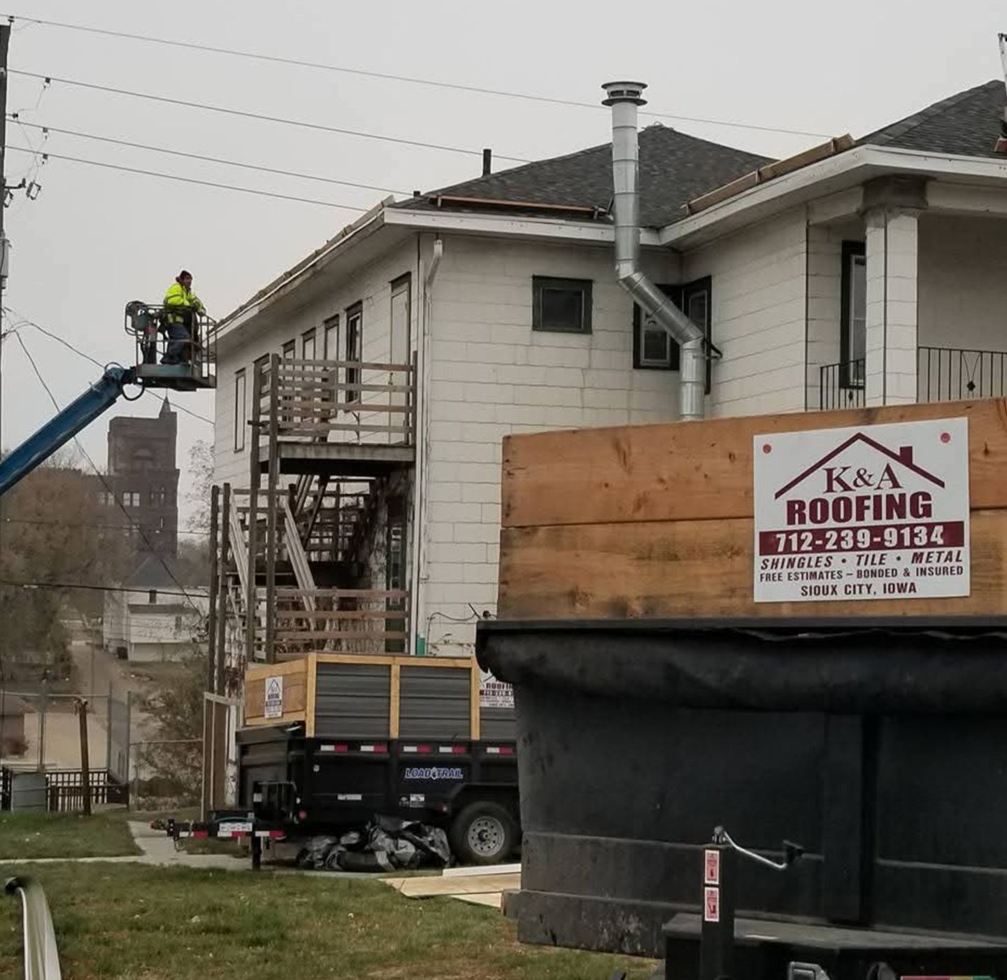 Roofing workers on a house beside a dumpster and lift, with K&A Roofing sign visible.