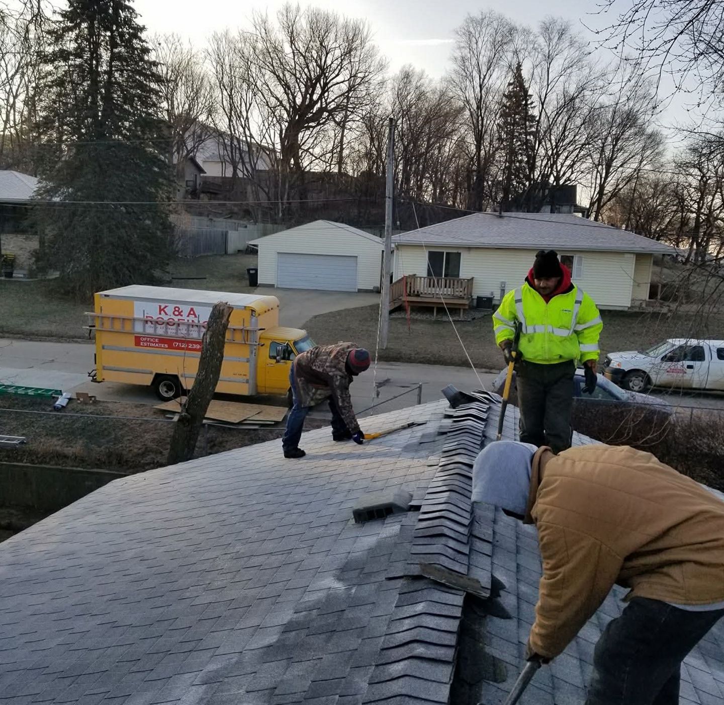 Workers on a rooftop installing shingles beside a yellow dumpster in a residential neighborhood.