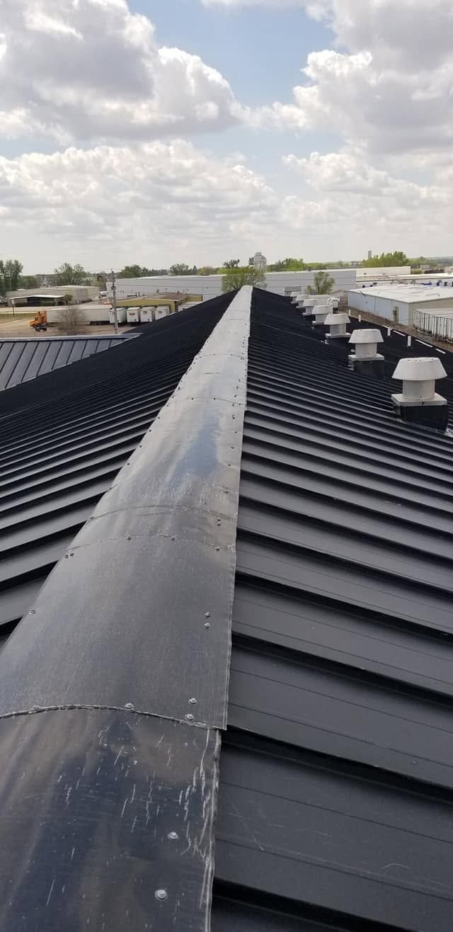 Rooftop with a long black metal ridge vent under a cloudy sky, surrounded by corrugated roofs