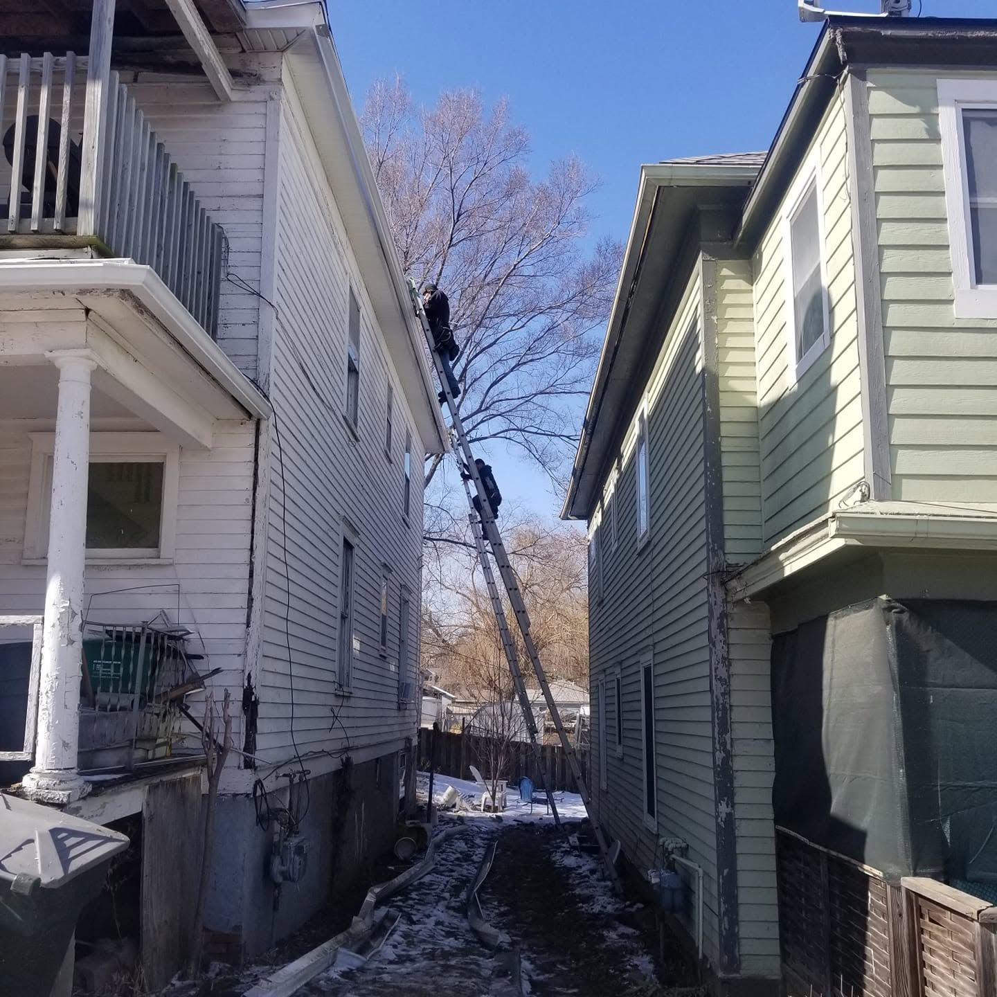 Narrow snowy alley between two houses with a tree at the end under a clear blue sky