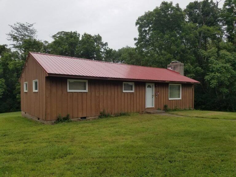 A brown house with a red roof is in the middle of a grassy field