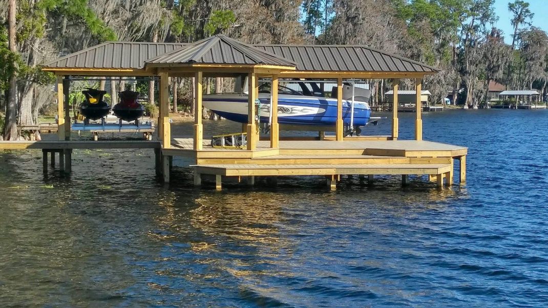 Wooden boat dock with two canopies and two boats on a lake, surrounded by trees.