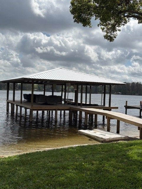 A boat dock with a white metal roof stands over water under a cloudy sky, connected to a grassy shore by a wooden walkway.