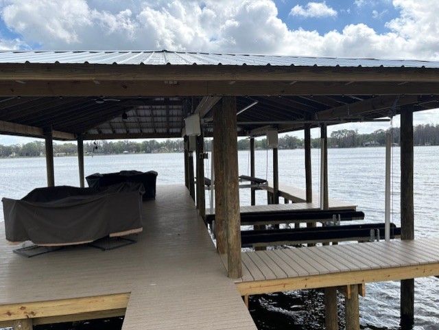A wooden boat dock with a metal roof over water, featuring covered lounge furniture and empty boat slips.