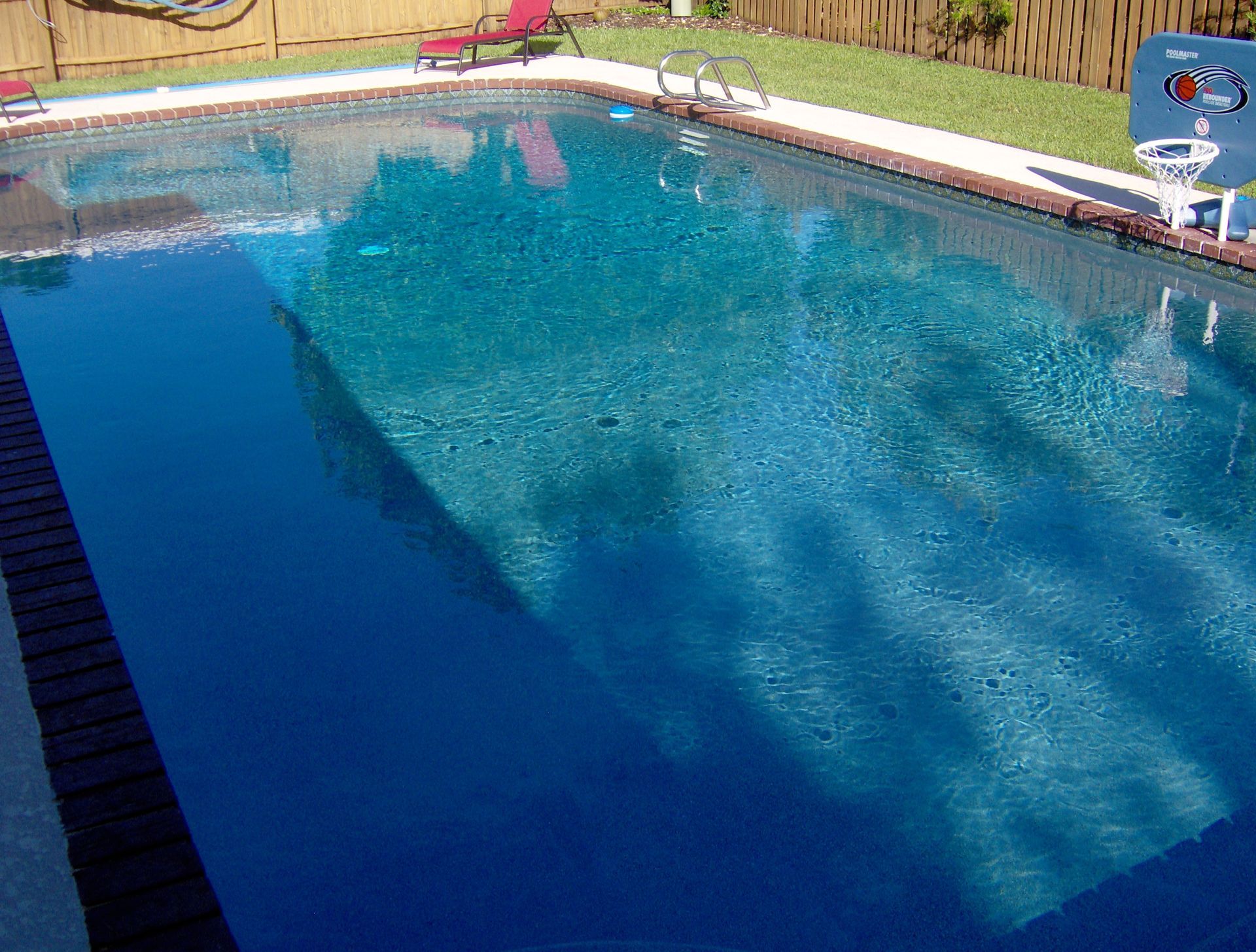 Backyard swimming pool with blue water, deck, and fence in sunlight.