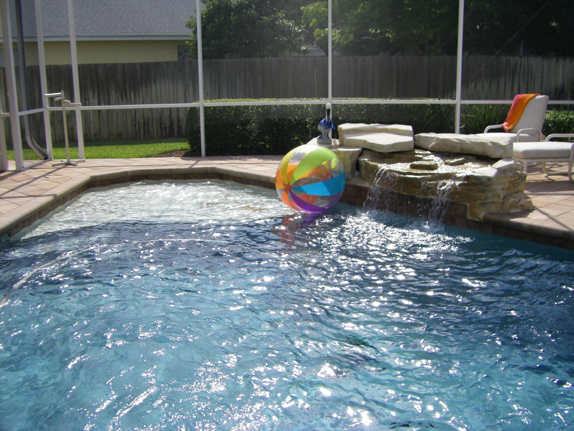 Colorful beach ball floating in a backyard swimming pool beside a stone waterfall and lounge chair