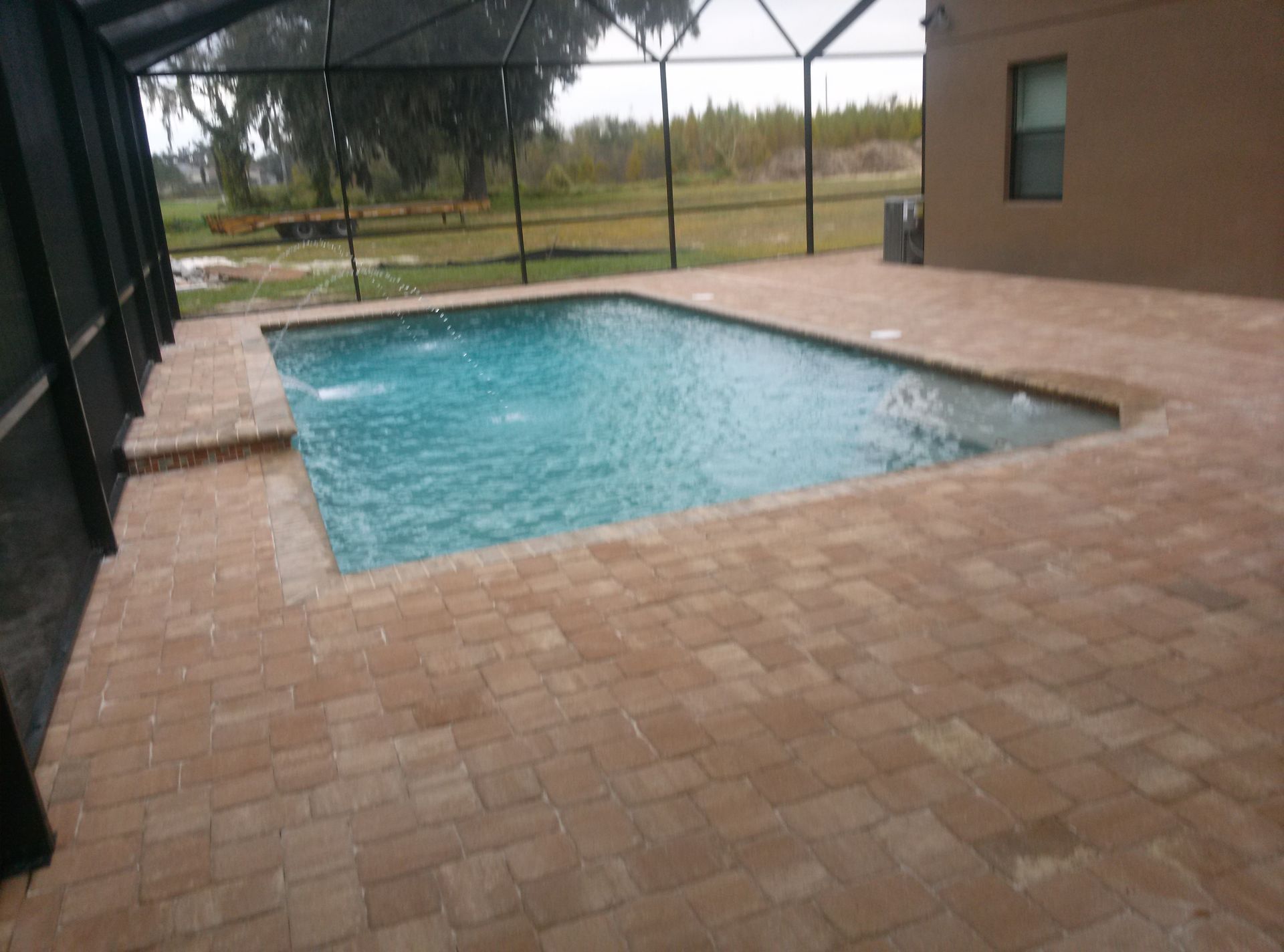 Covered backyard pool with blue water and tan paver patio beside a house