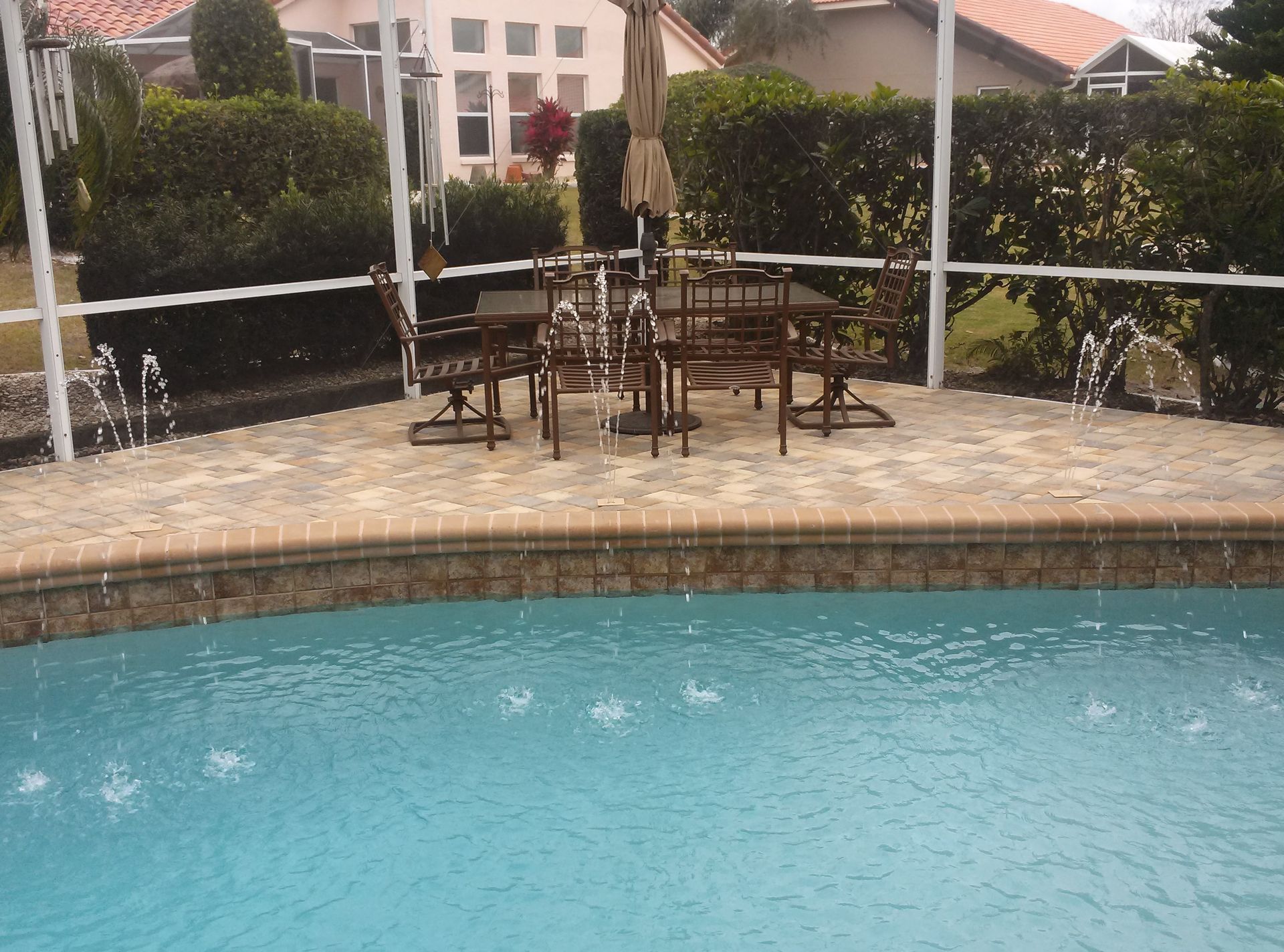 Backyard pool with stone deck, patio table and chairs, and hedge-lined fence under a shaded umbrella