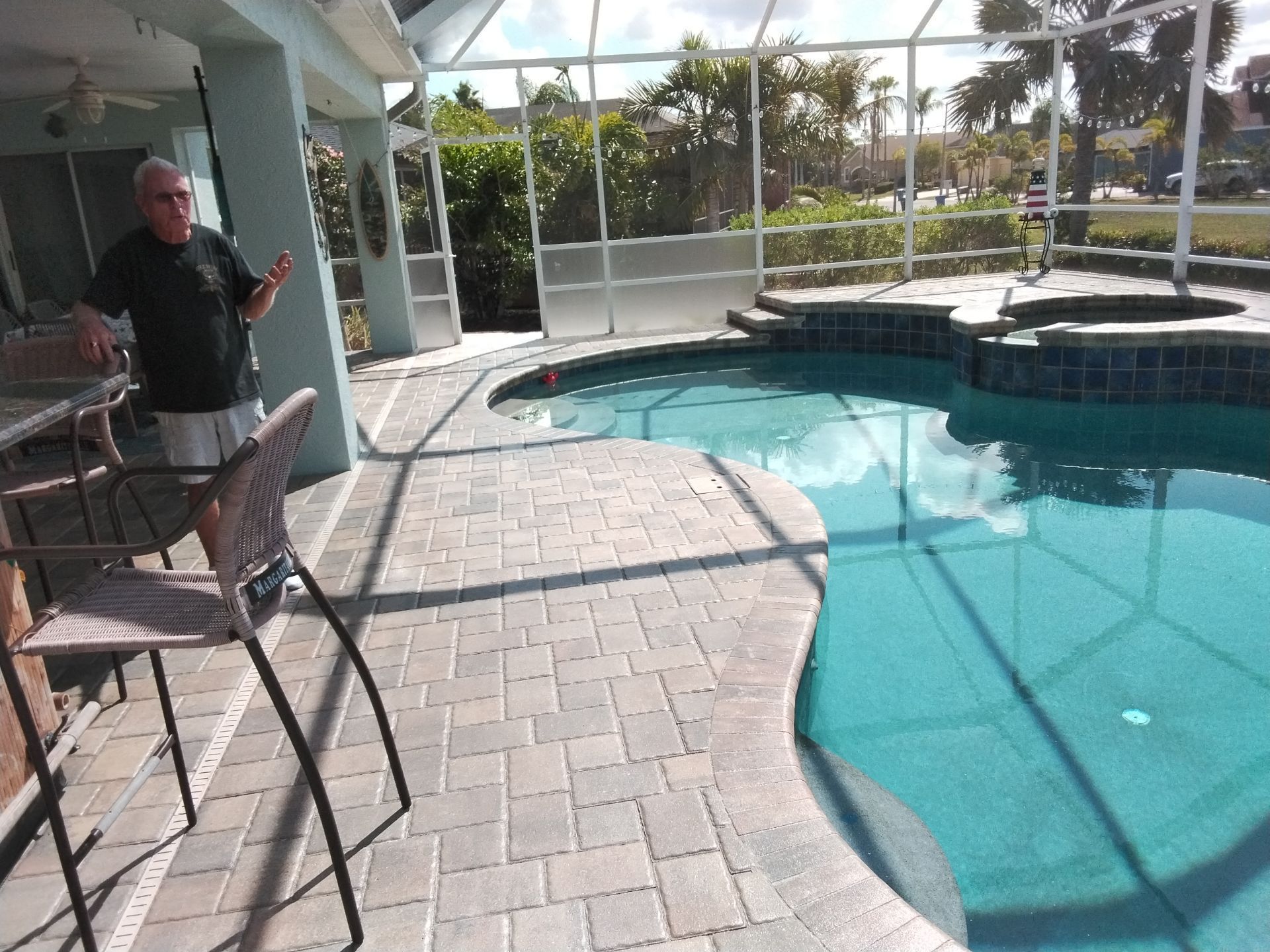 Man standing beside a patio table near a backyard pool and screened enclosure.