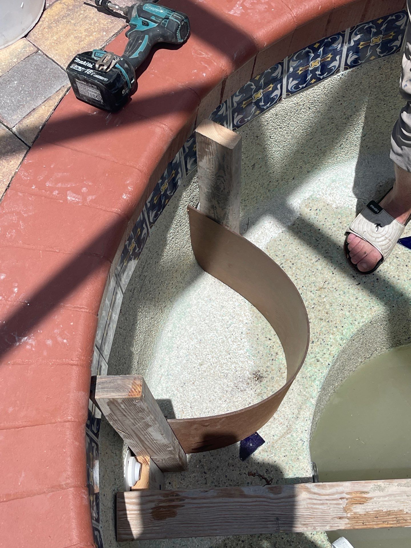 Construction materials around a curved pool drain, with a tool and workers’ feet visible on tiled deck.