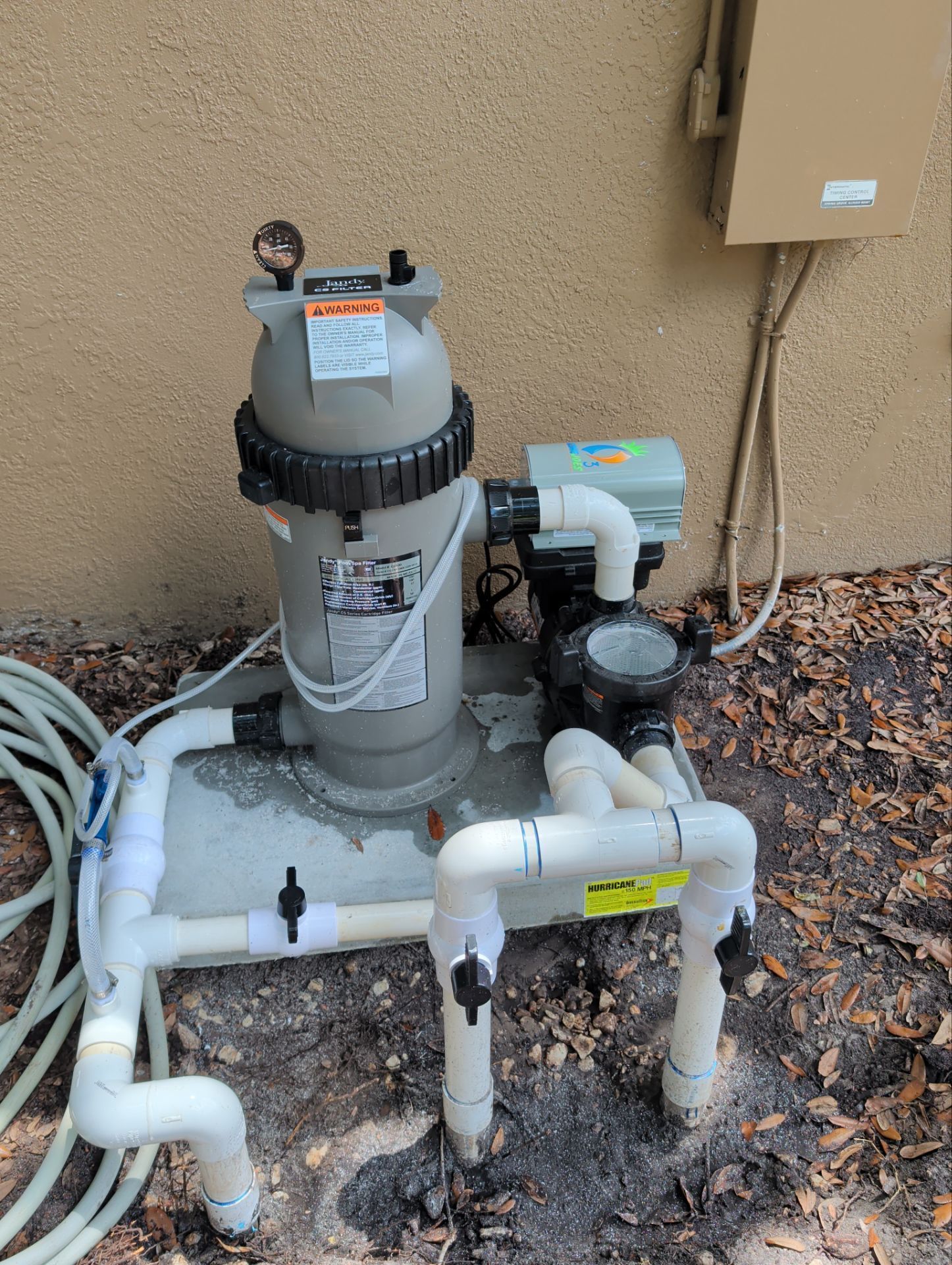 Pool pump and filter system with white pipes beside a tan wall and dirt ground.