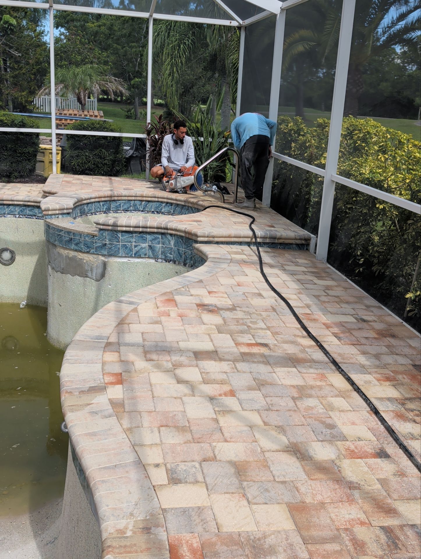 Workers pressure-wash a stone poolside walkway beside a covered patio and pool.