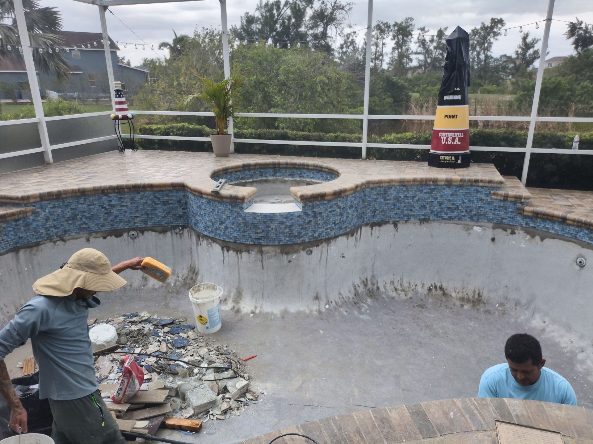 Workers cleaning a drained swimming pool with debris on the bottom and a small spa at the far end.