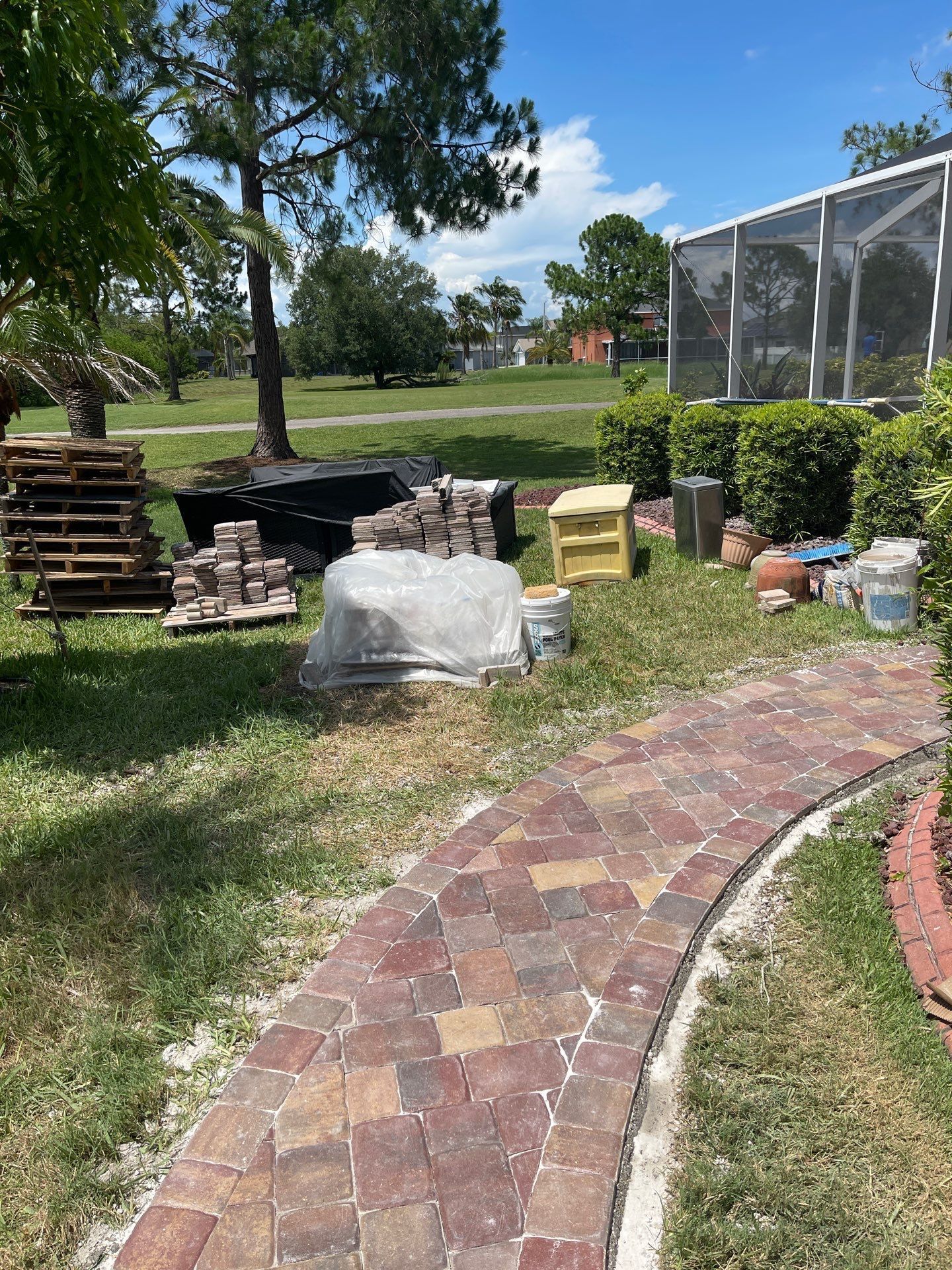 Backyard with stacked firewood, covered materials, and a curved brick path beside a screened enclosure.