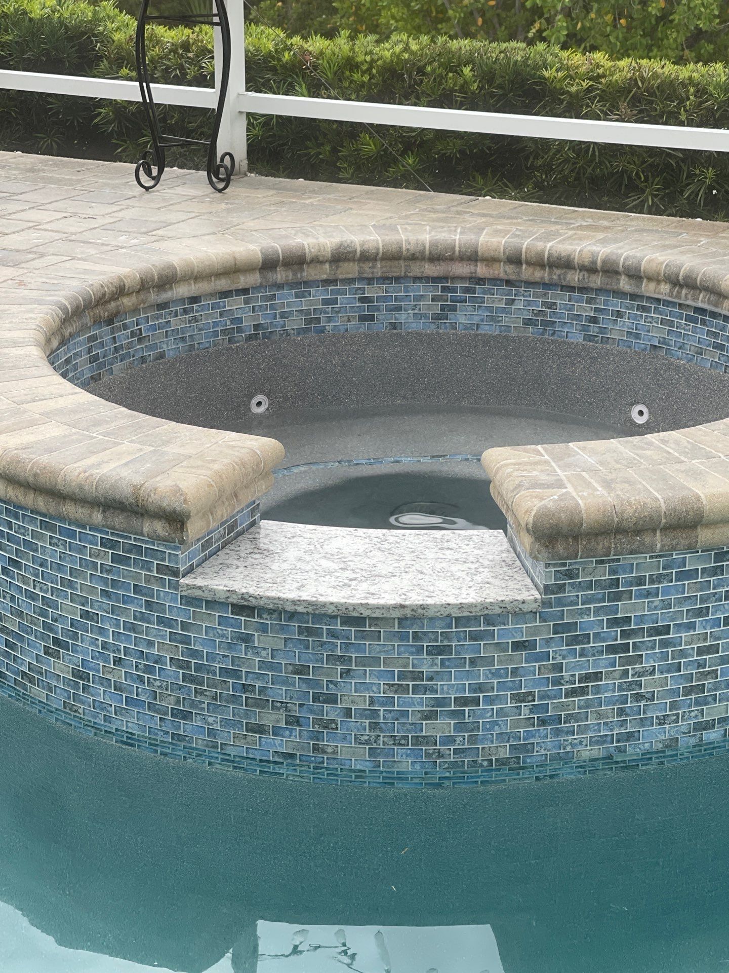 Small tiled pool spa with blue mosaic walls and beige stone coping beside a larger pool.