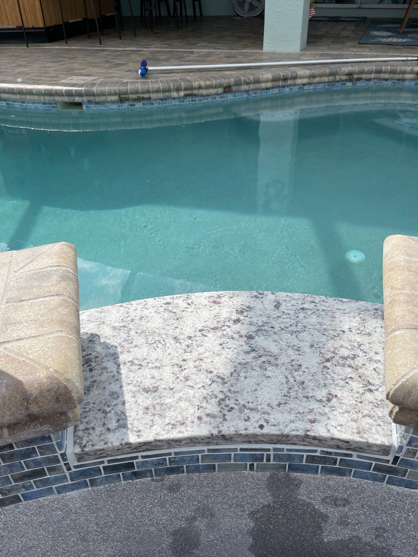 Backyard swimming pool with stone steps, tan cushions, and clear blue water.