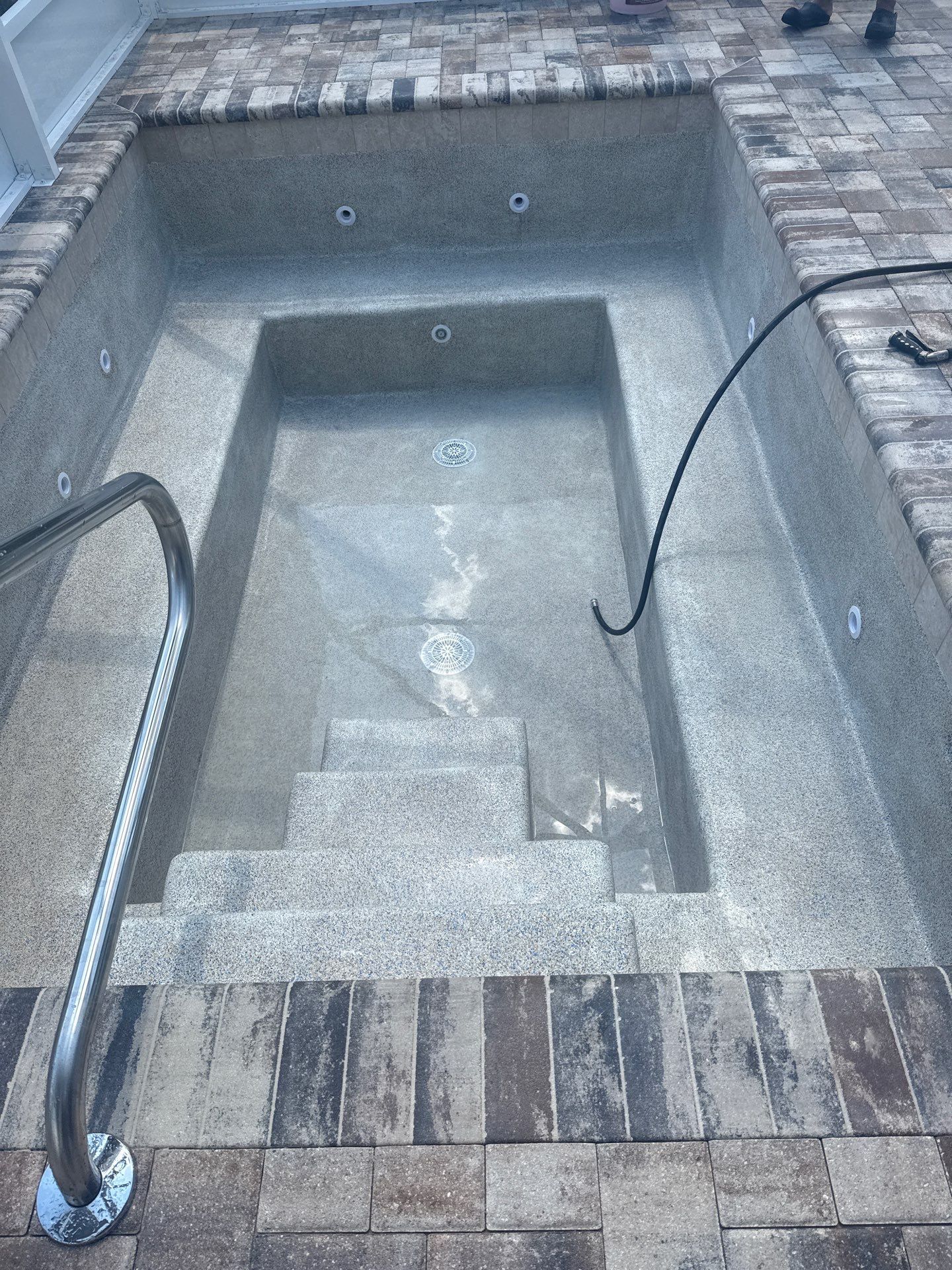 Empty concrete pool with steps and two black hoses, viewed from above.
