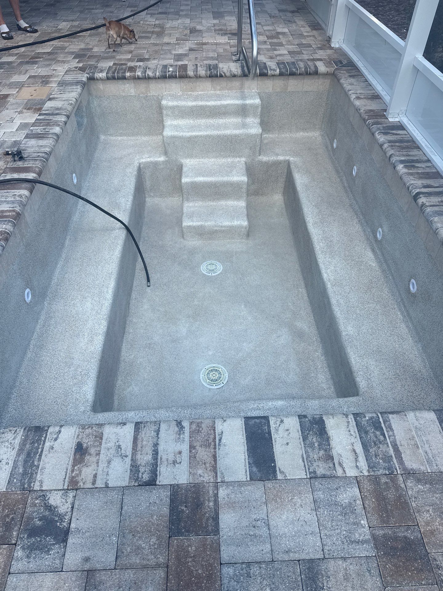 Empty rectangular in-ground pool with steps, surrounded by brick pavers and a pool hose.
