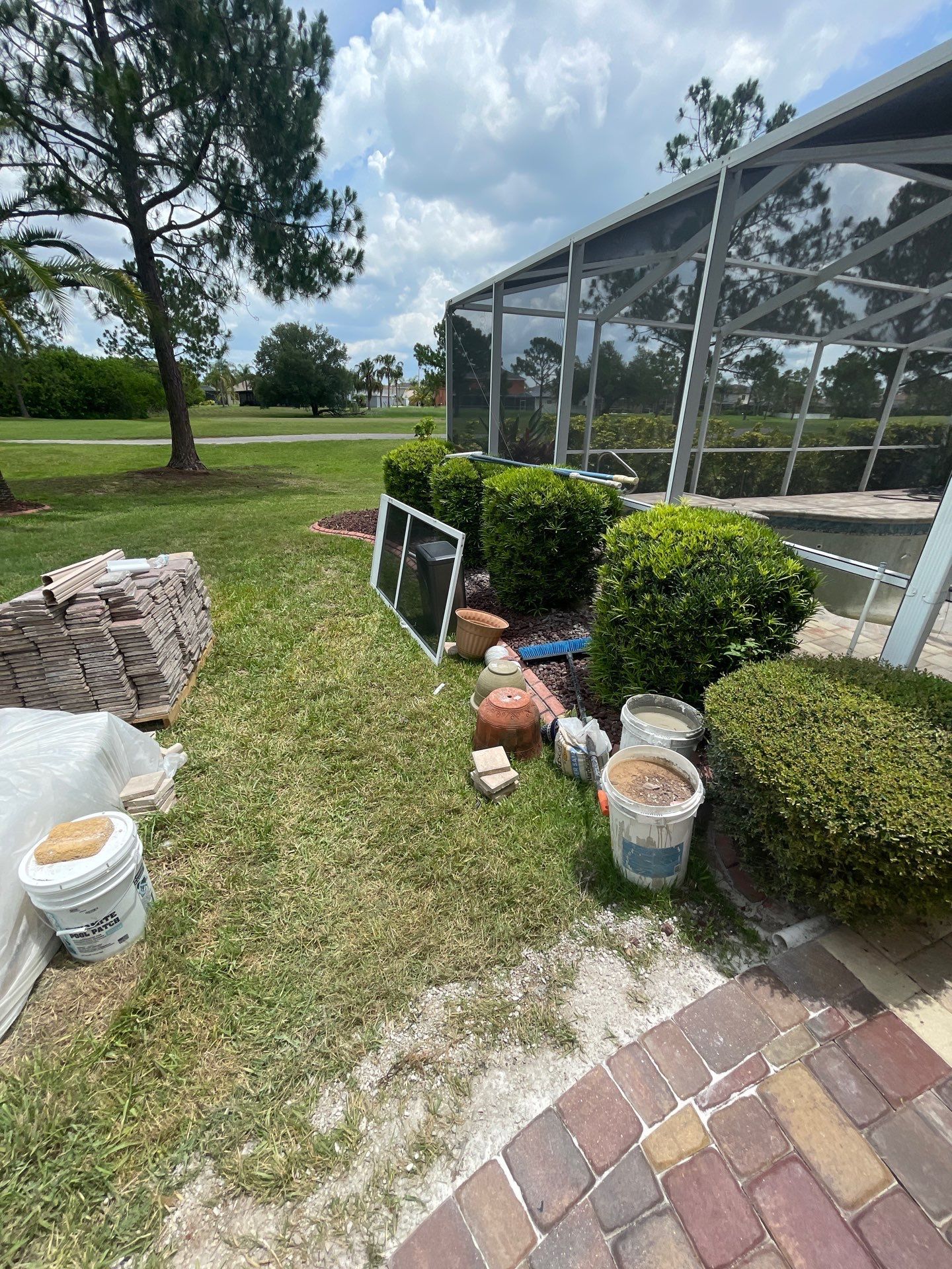 Backyard patio with potted plants, bushes, and stacked stones beside a screened enclosure.
