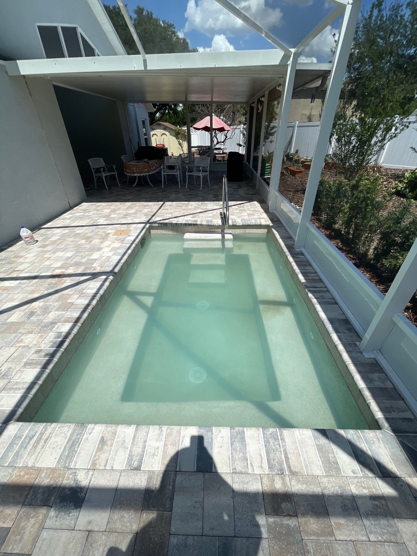 Small rectangular pool under a covered patio, with glass railings and tiled deck in bright sunlight.