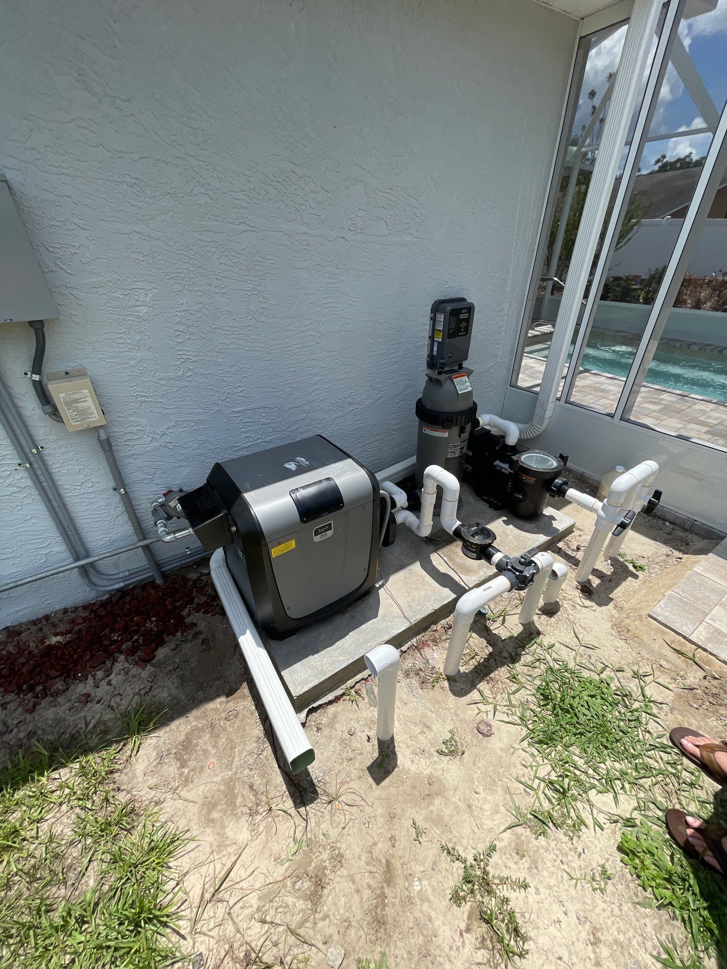 Pool equipment beside a house wall: gray pump, black filter, and white pipes on a concrete pad.