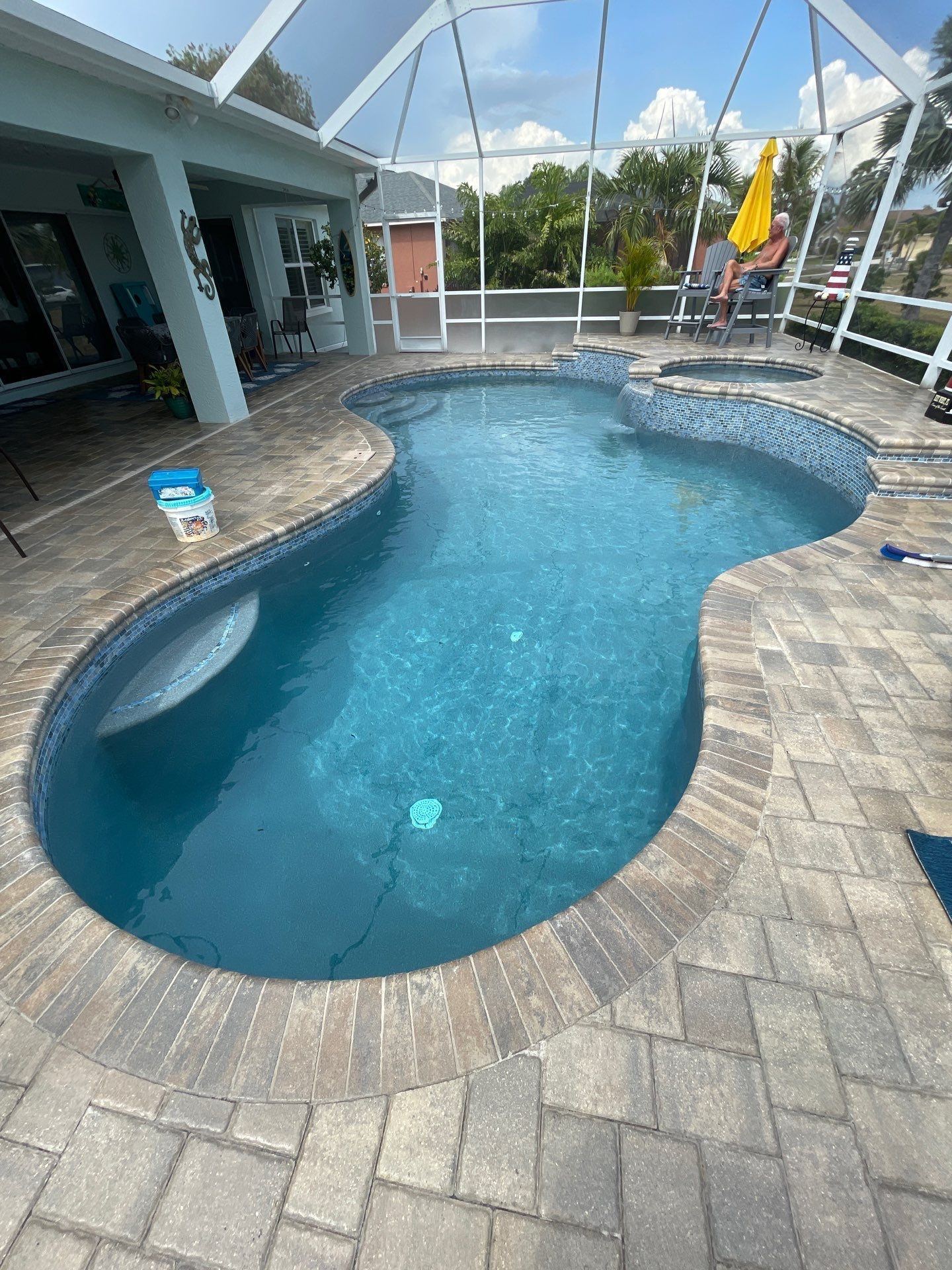 Curved backyard swimming pool with blue water, stone pavers, and patio seating under a covered lanai.