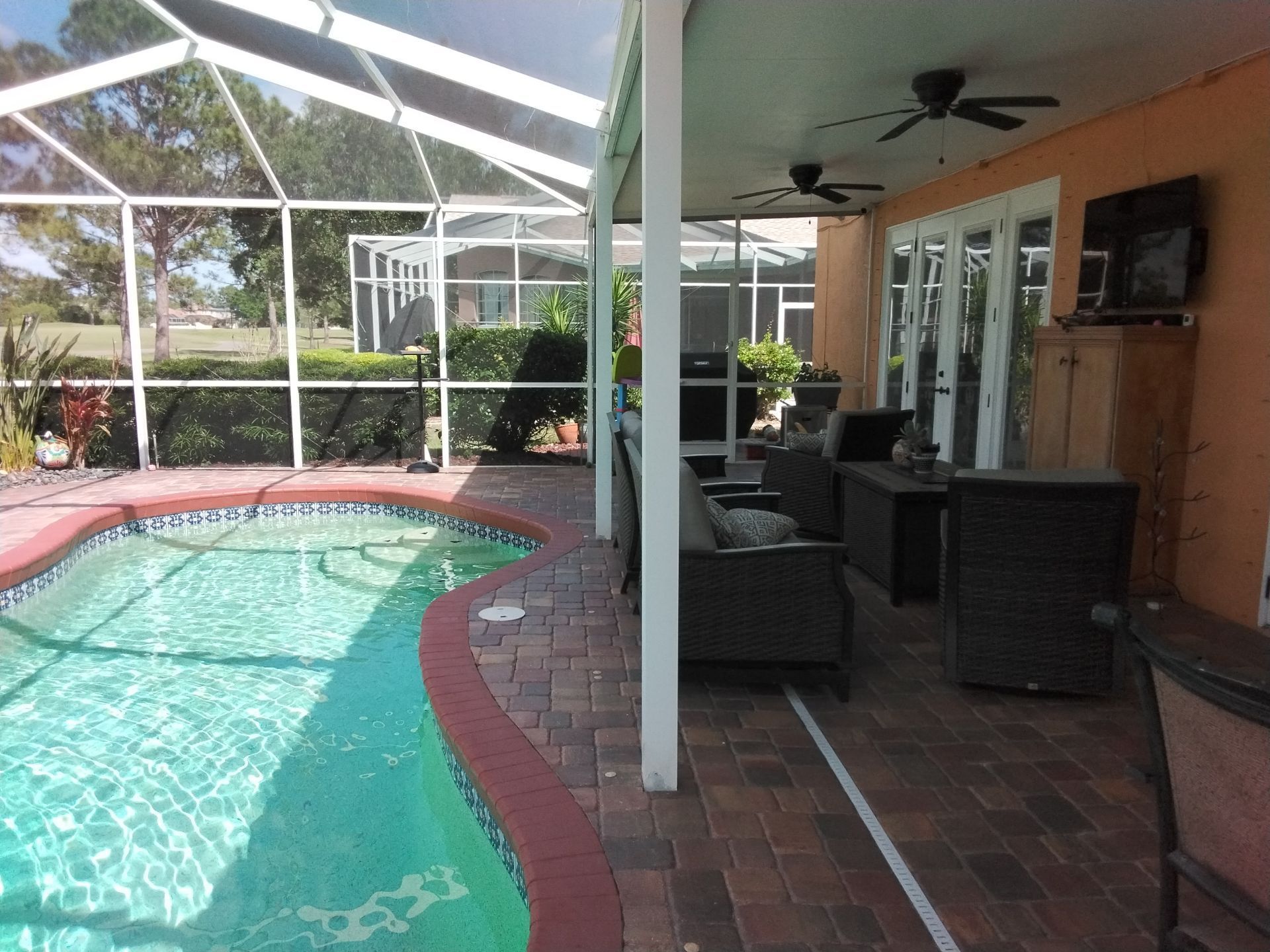 Covered patio with wicker chairs beside a screened-in swimming pool and brick pavers.