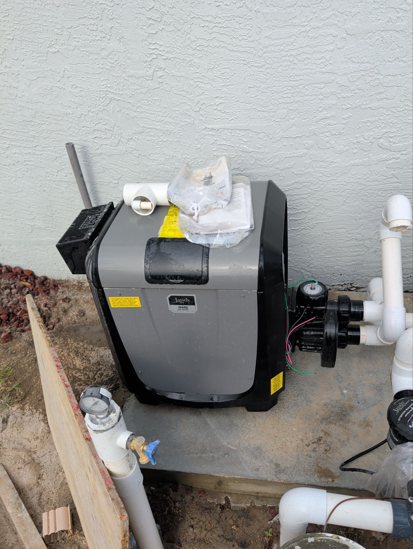 Outdoor pool equipment with gray heater unit, black pump, and white PVC pipes beside a wall.