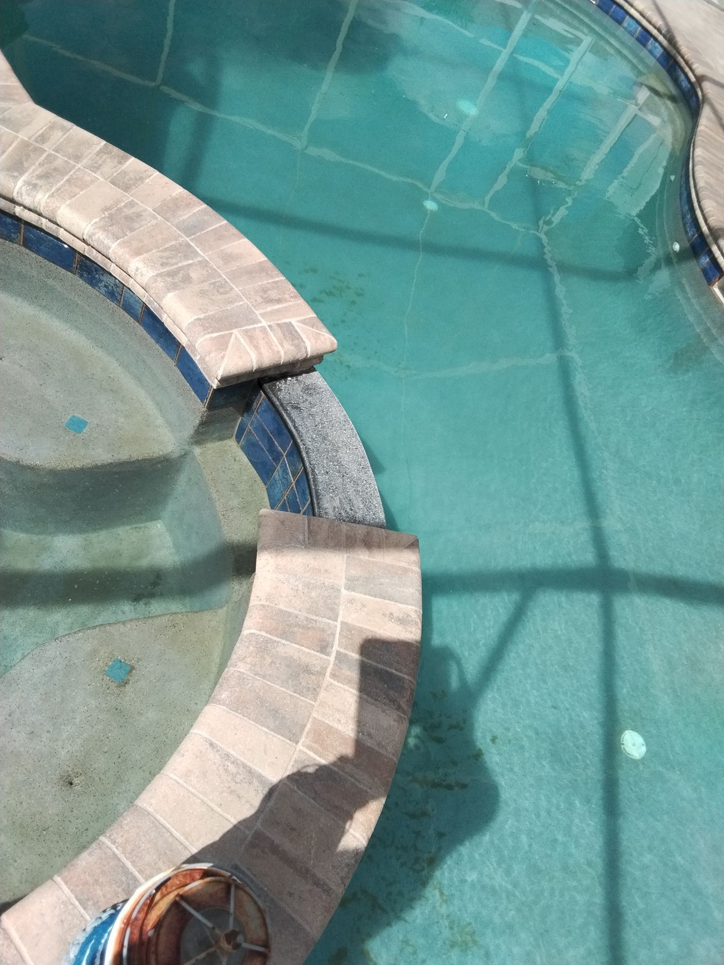 Curved stone hot tub beside a turquoise swimming pool with a tiled deck and shadows.